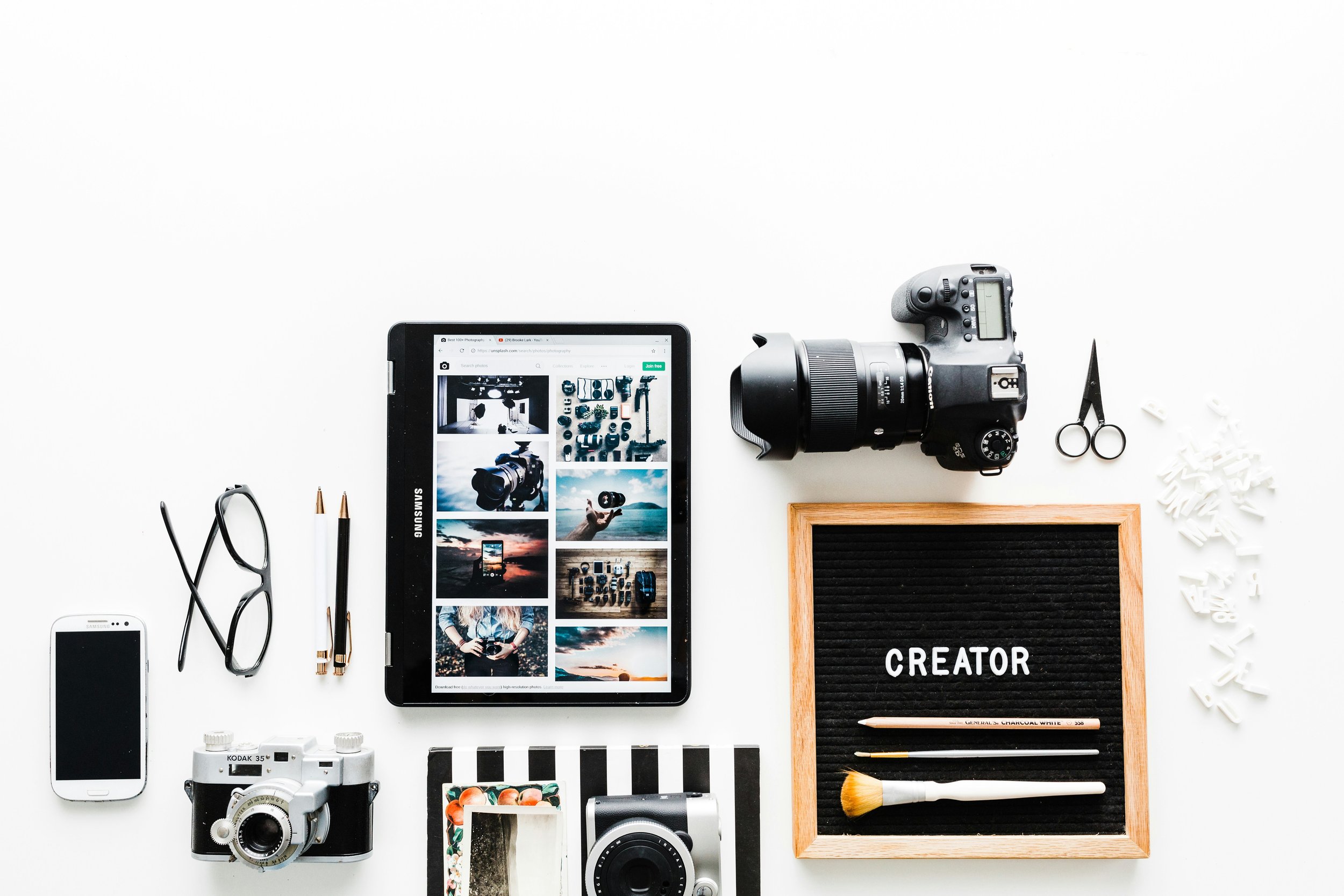 Cameras, a tablet, phone, glasses, and a letter board spelling “Creator” arranged on a white desk.