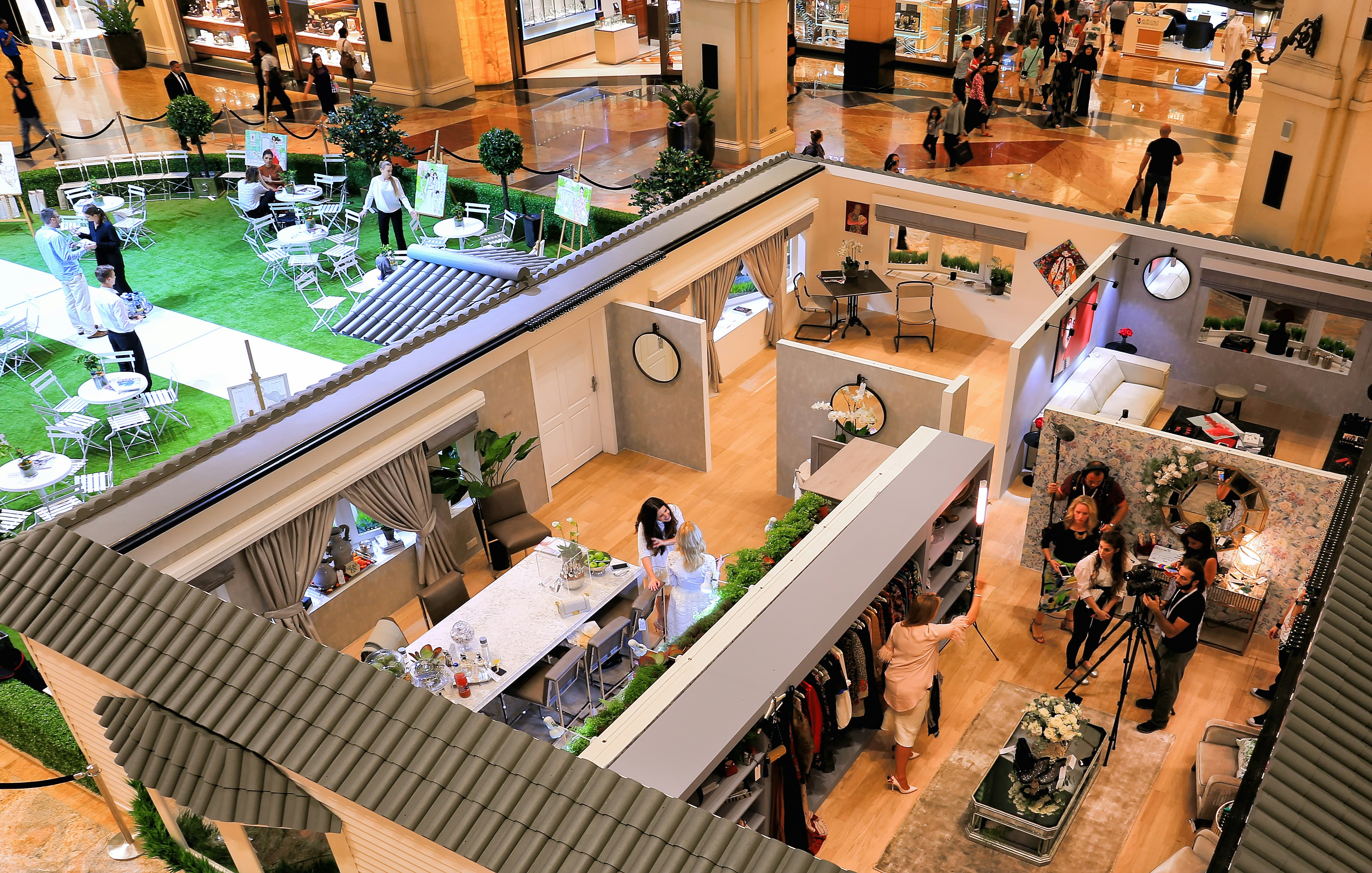 A large indoor brand event includes staged rooms, tables, displays, camera crews, and groups of attendees moving through the space.