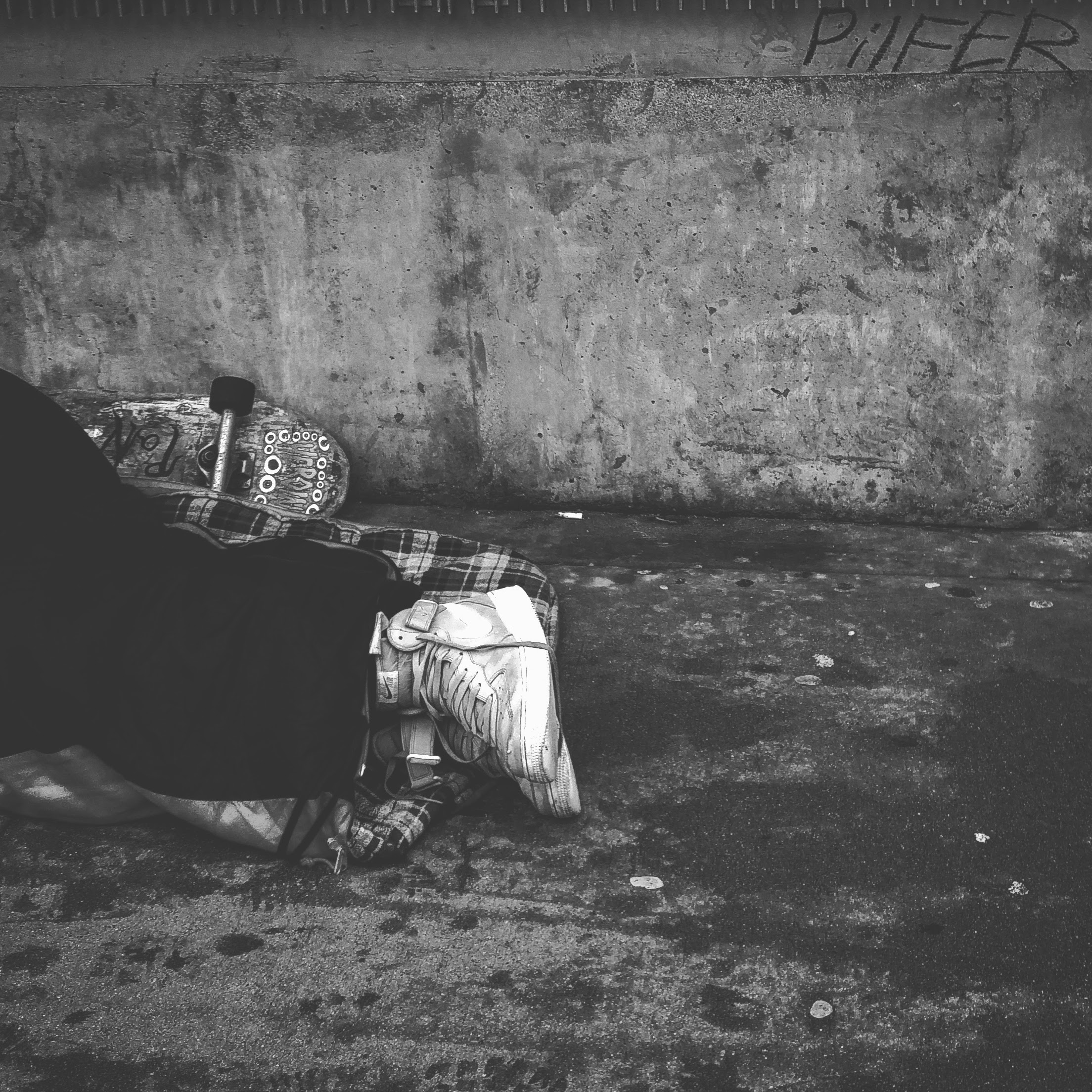 A person laying down on the street on top of a blanket. Their legs, Nike shoes and skateboard next to them.