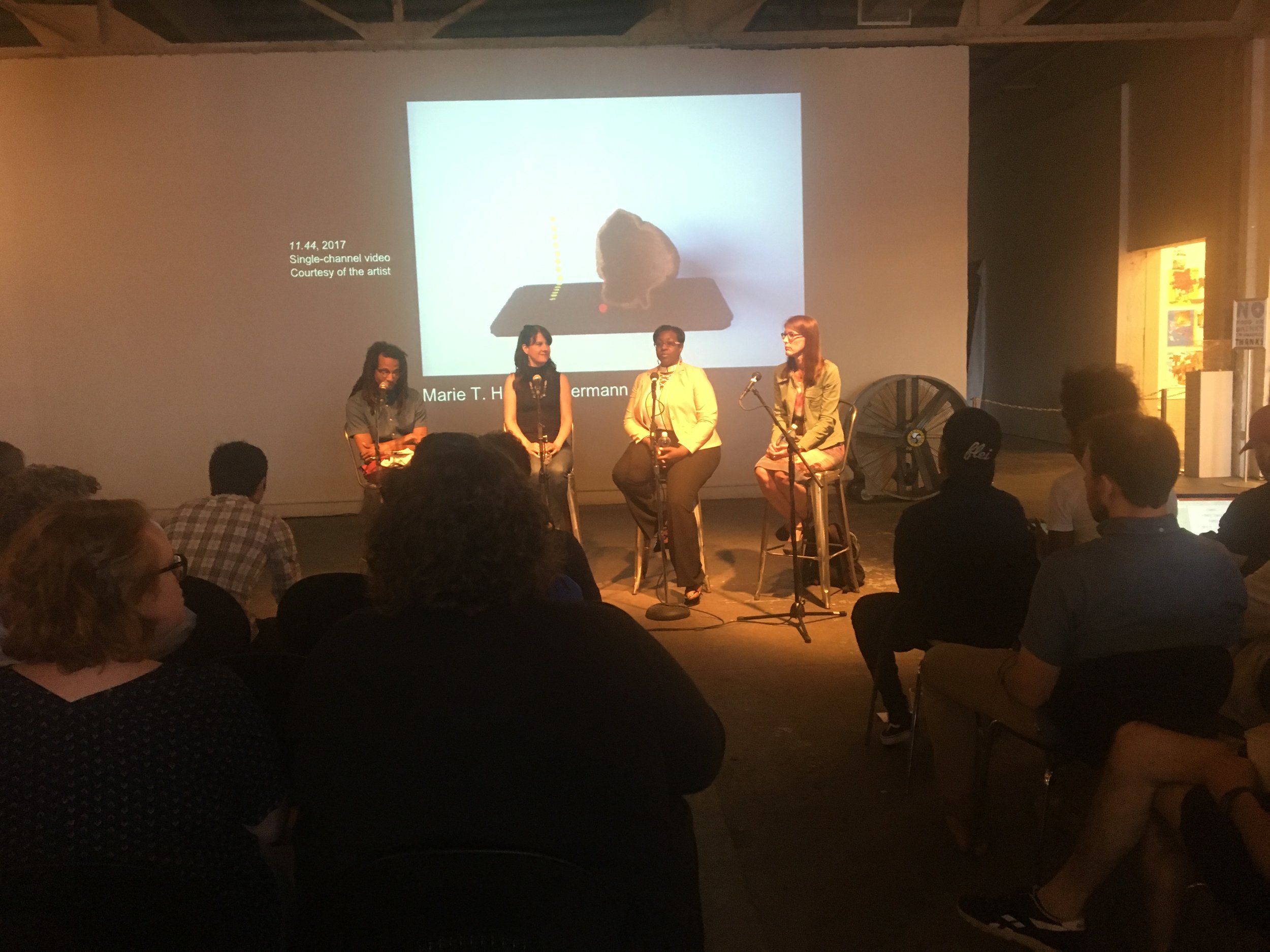 The three panelists and the moderator sitting on stools in front of an audioence at the MOCAD.