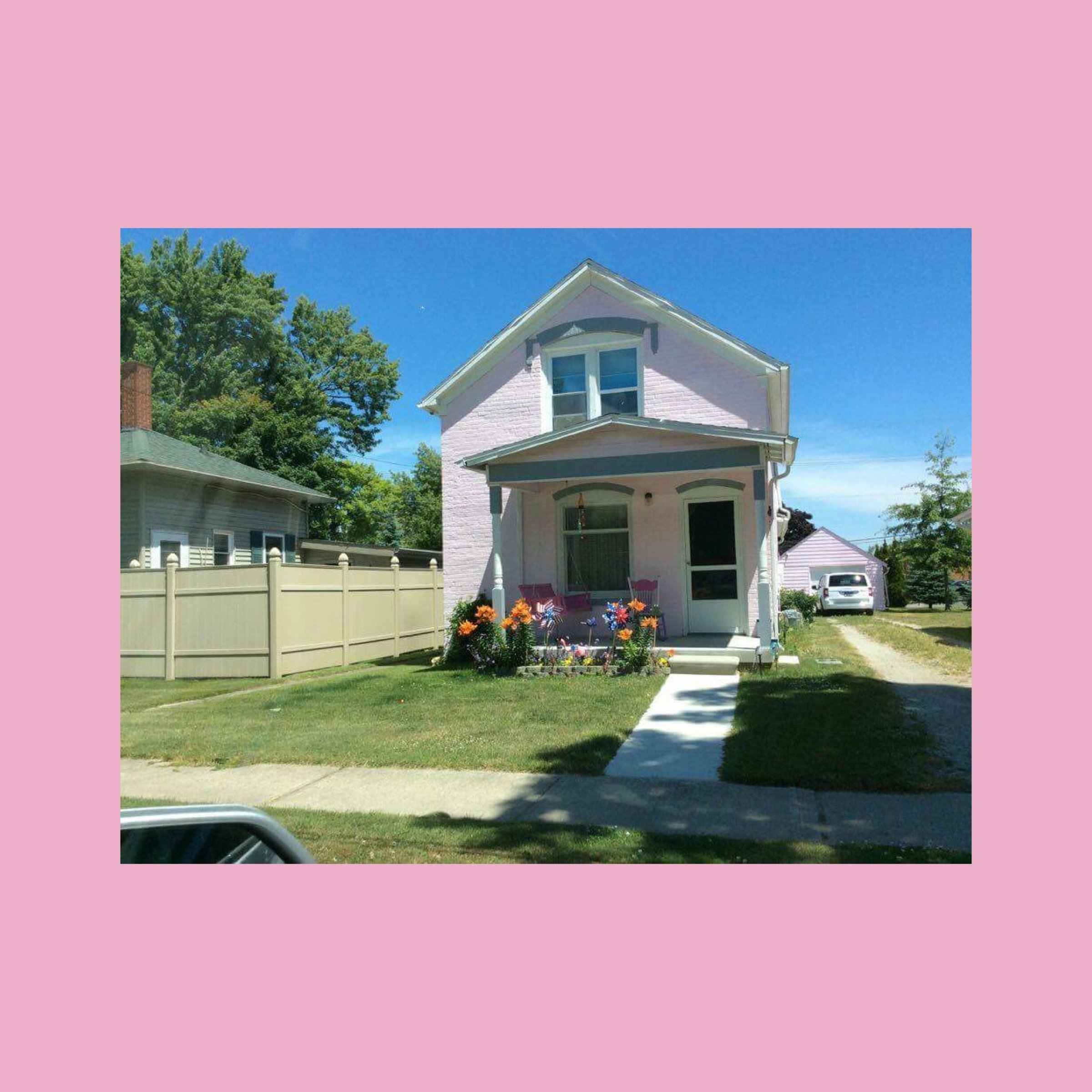 A pale pink two-story house with flowers along the front porch on a sunny day.