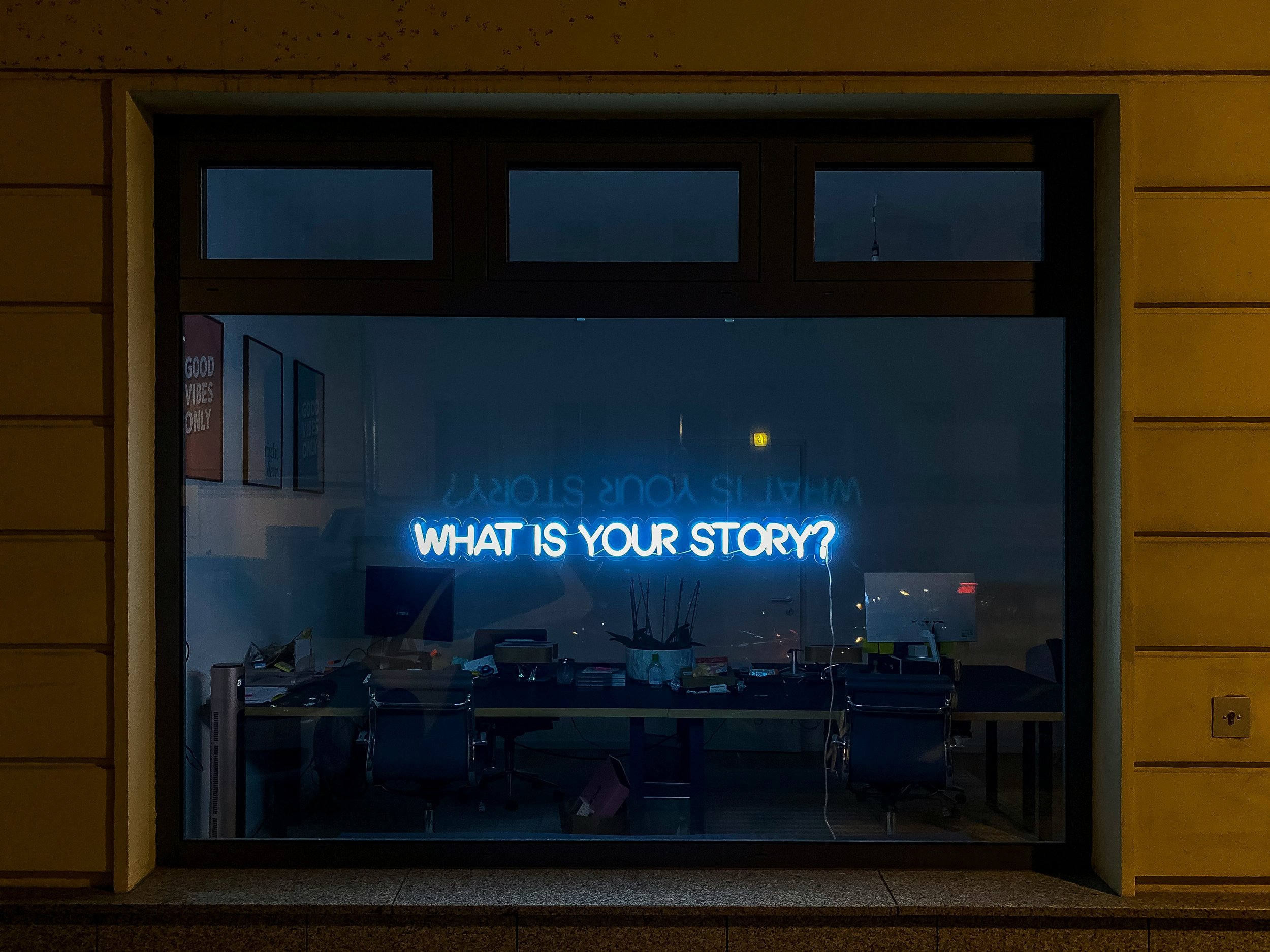 A neon sign reading “What is your story?” glows inside a dark office window.